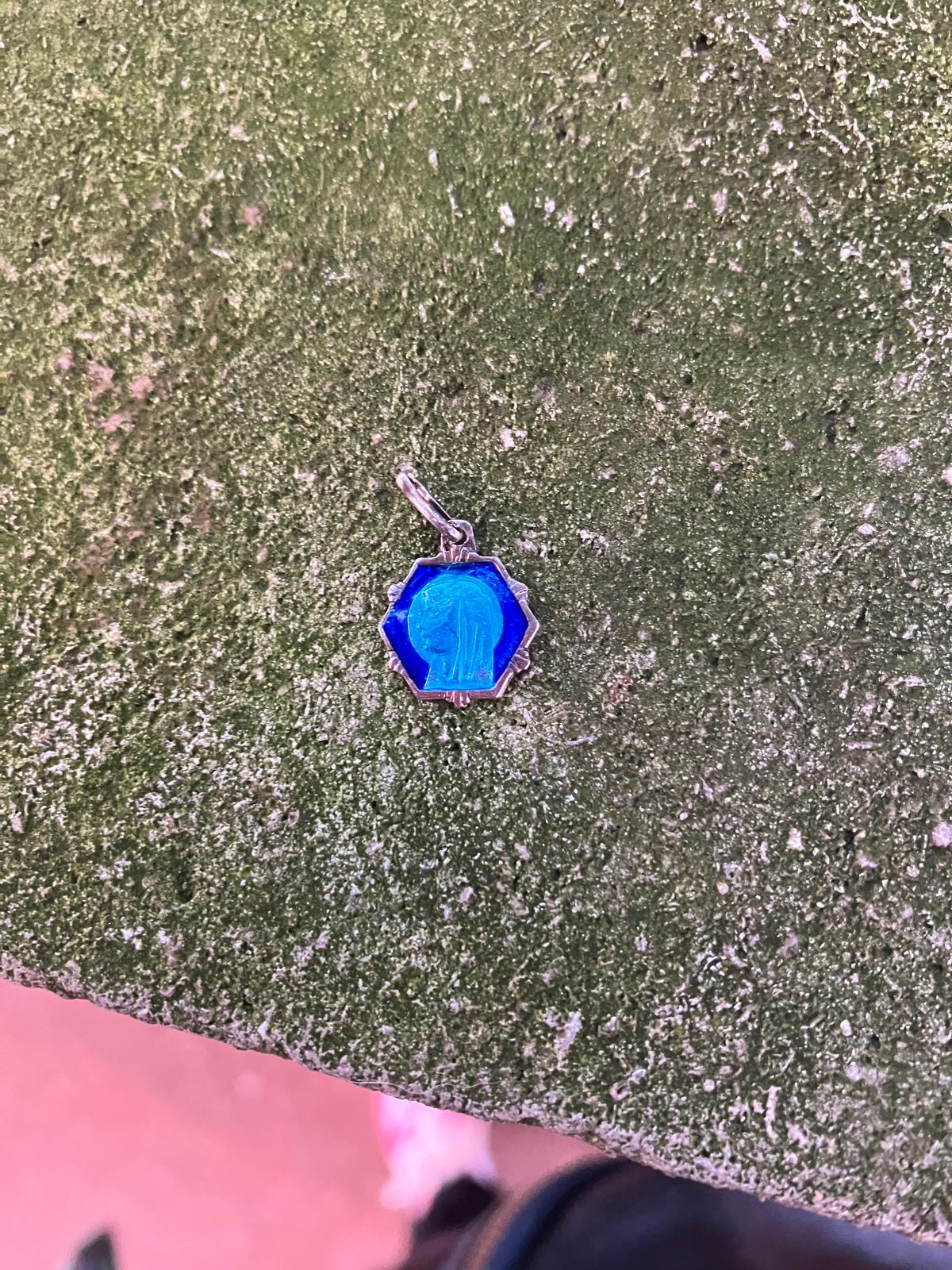 Médaille religieuse bleue en émail, de forme hexagonale, gravée d'une silhouette de la Vierge, posée sur une pierre moussue